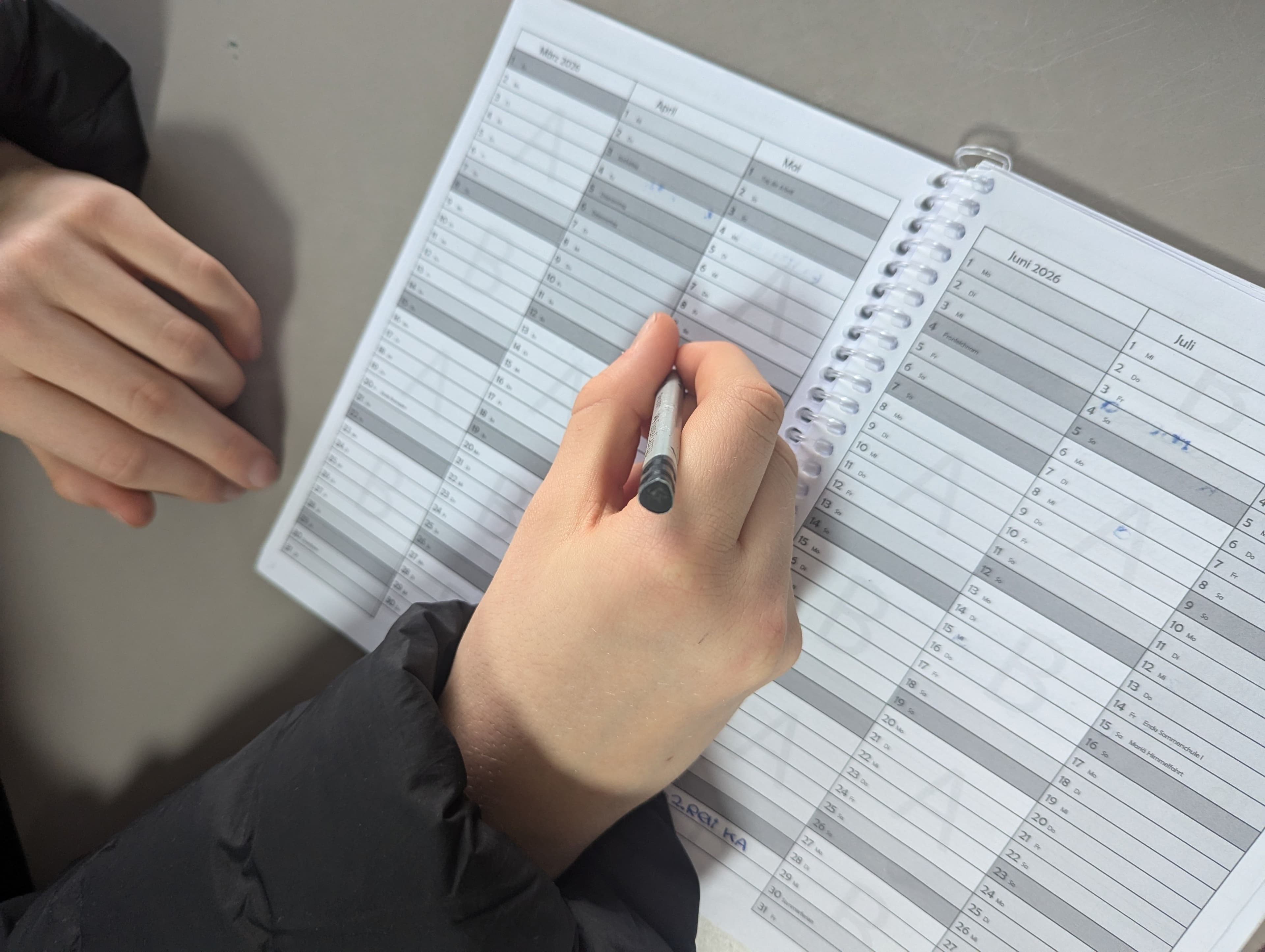 A hand writes with a pen in an open spiral-bound calendar planner.