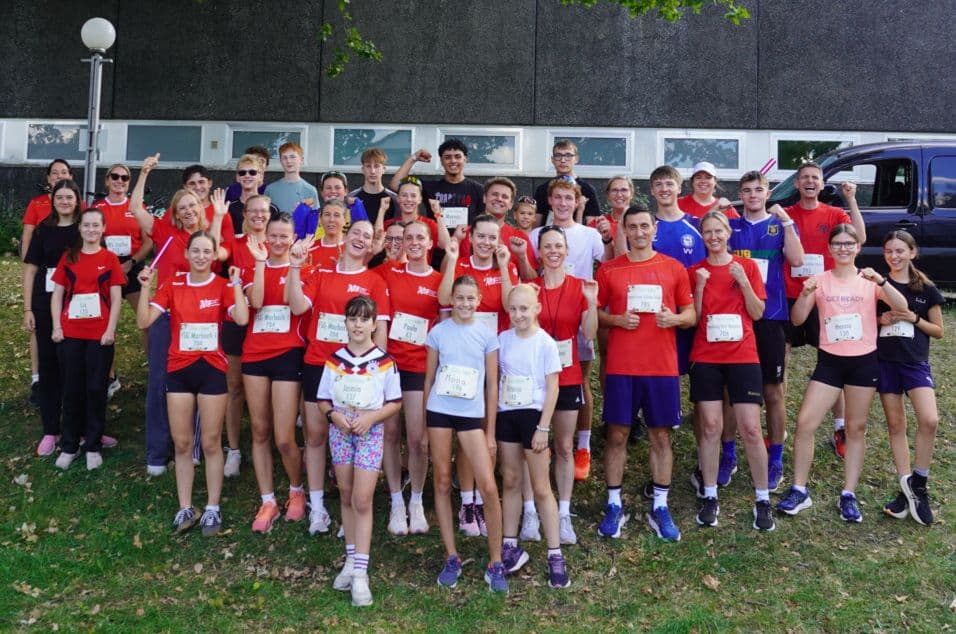 A large group of smiling people, many in red shirts with race bibs, pose outdoors on grass.