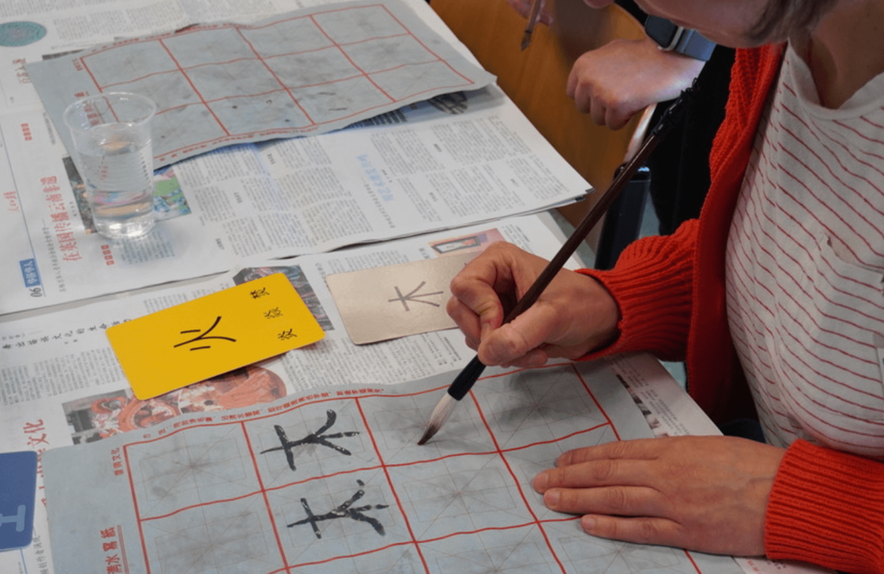 Hands practicing Chinese calligraphy with a brush on a gridded sheet of paper.
