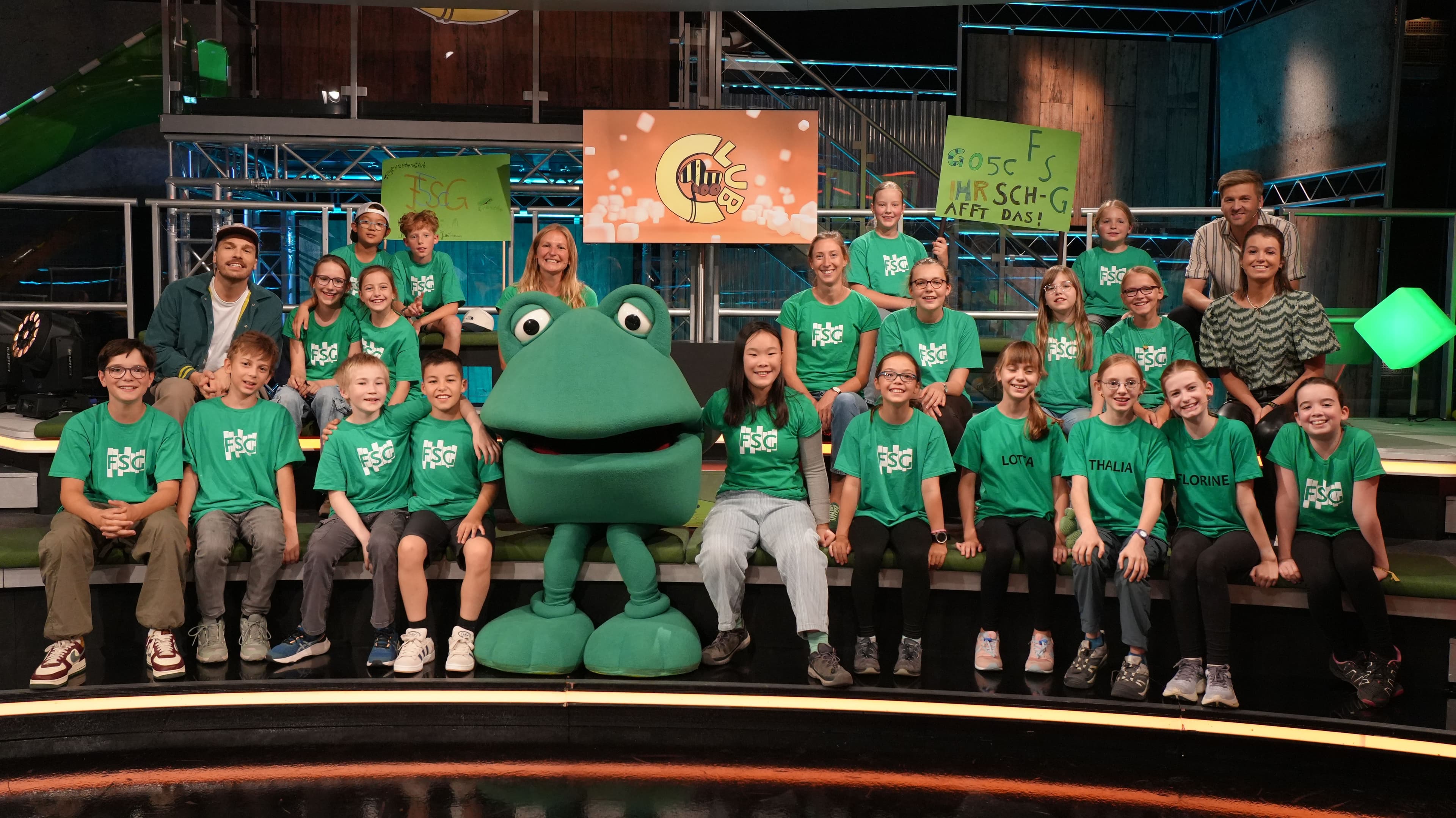 A group of children, two adults, and a green frog mascot in green shirts sit on a TV show set.