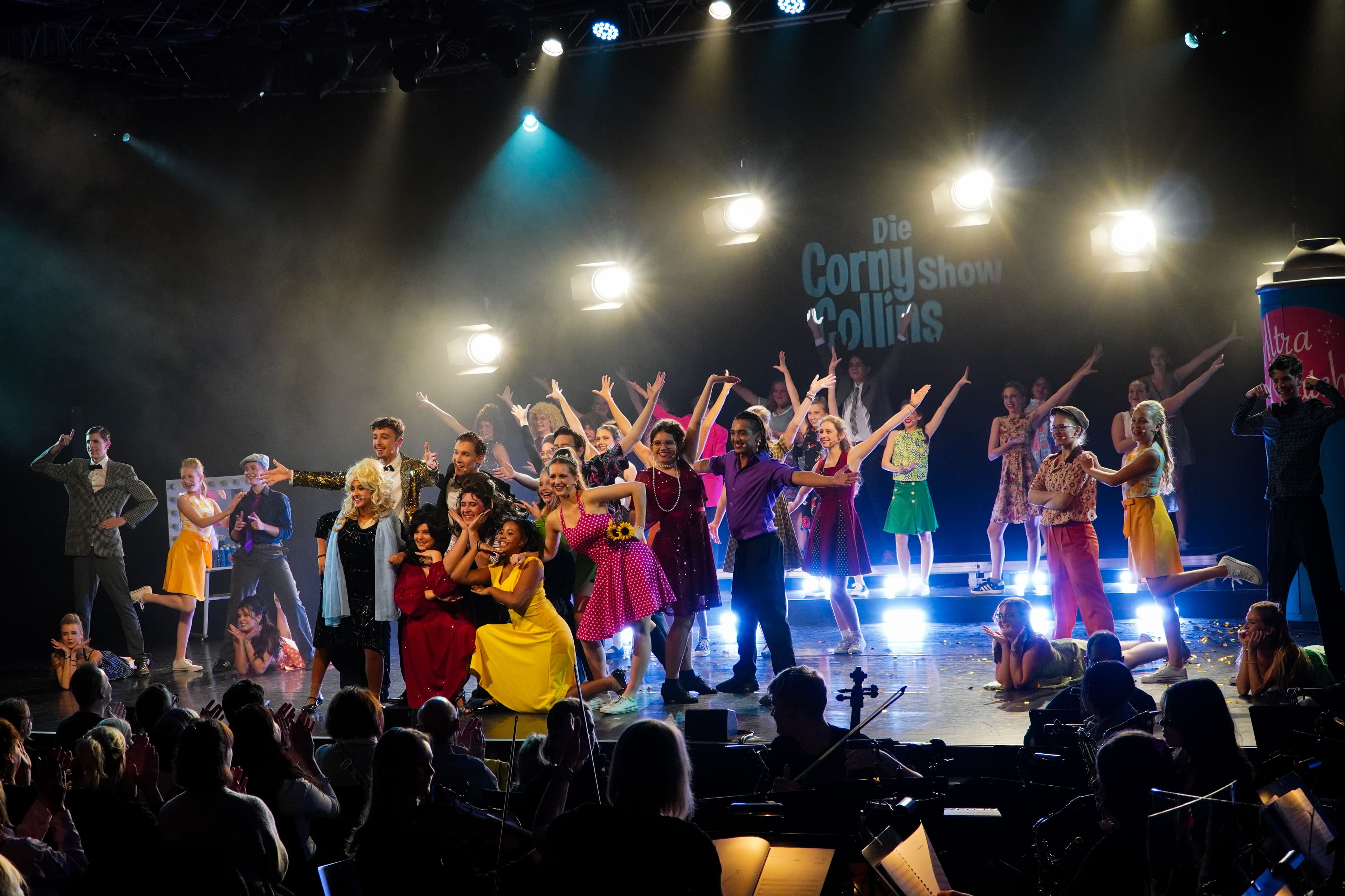 A large cast in colorful 1950s-style costumes poses on a brightly lit stage under a sign reading "Die Corny show Collins," with an audience applauding.