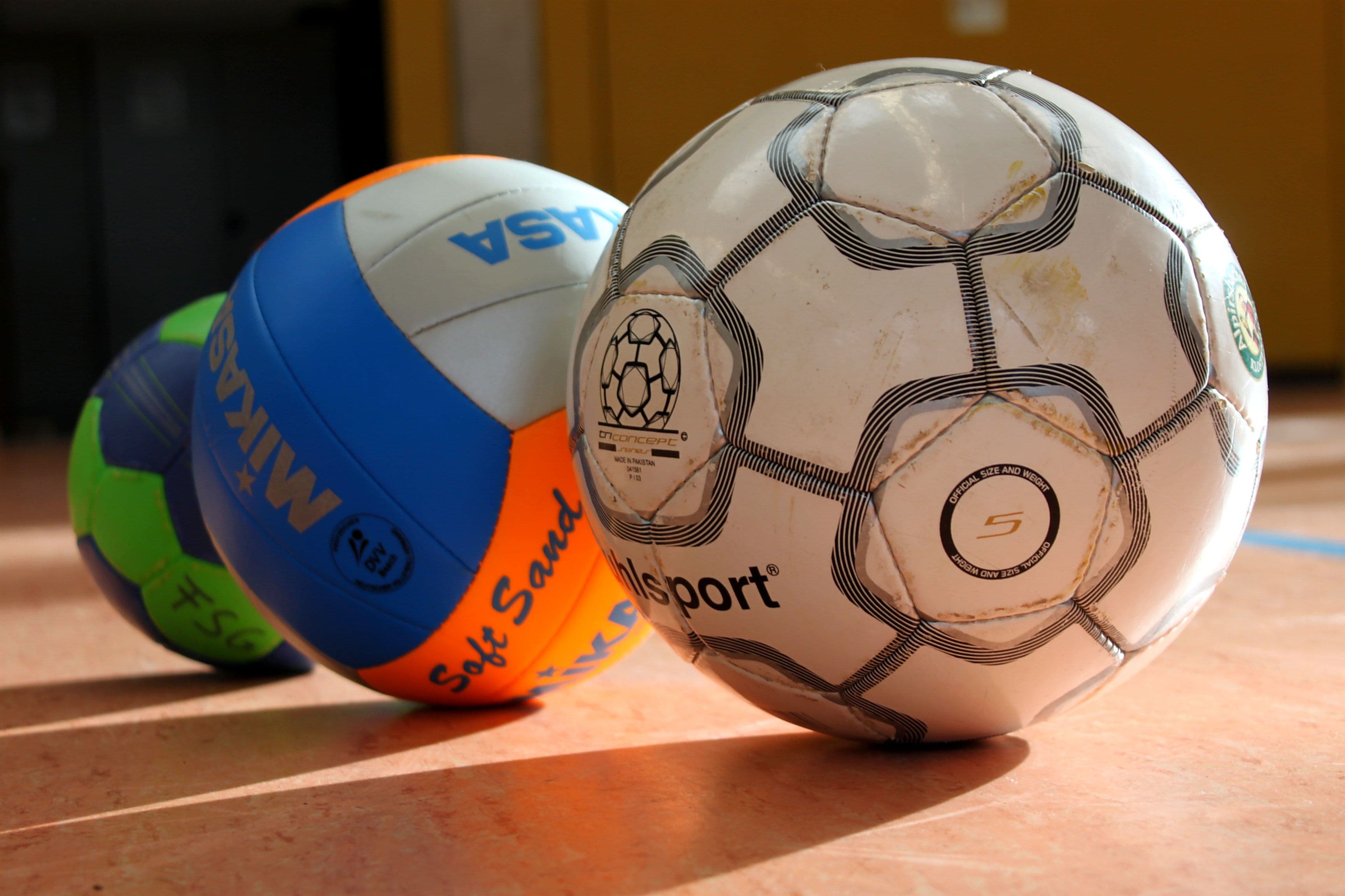 Ein Fußball, ein Volleyball, ein Handball in einer Reihe auf einem Hallenboden liegend.