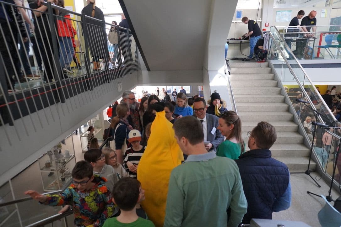 A person in a banana costume stands among a crowd in a multi-level building with stairs, while technicians operate equipment on an upper floor.