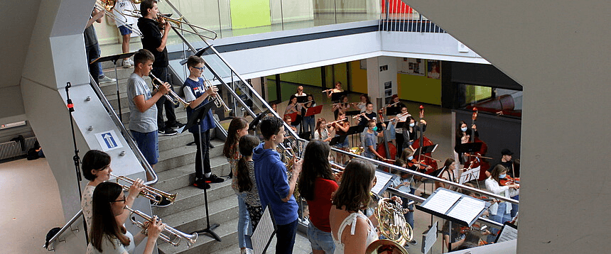 eine Gruppe von Menschen spielt ein Bläser- und Streicherkonzert im Schulhaus, auch auf der Treppe in der Aula