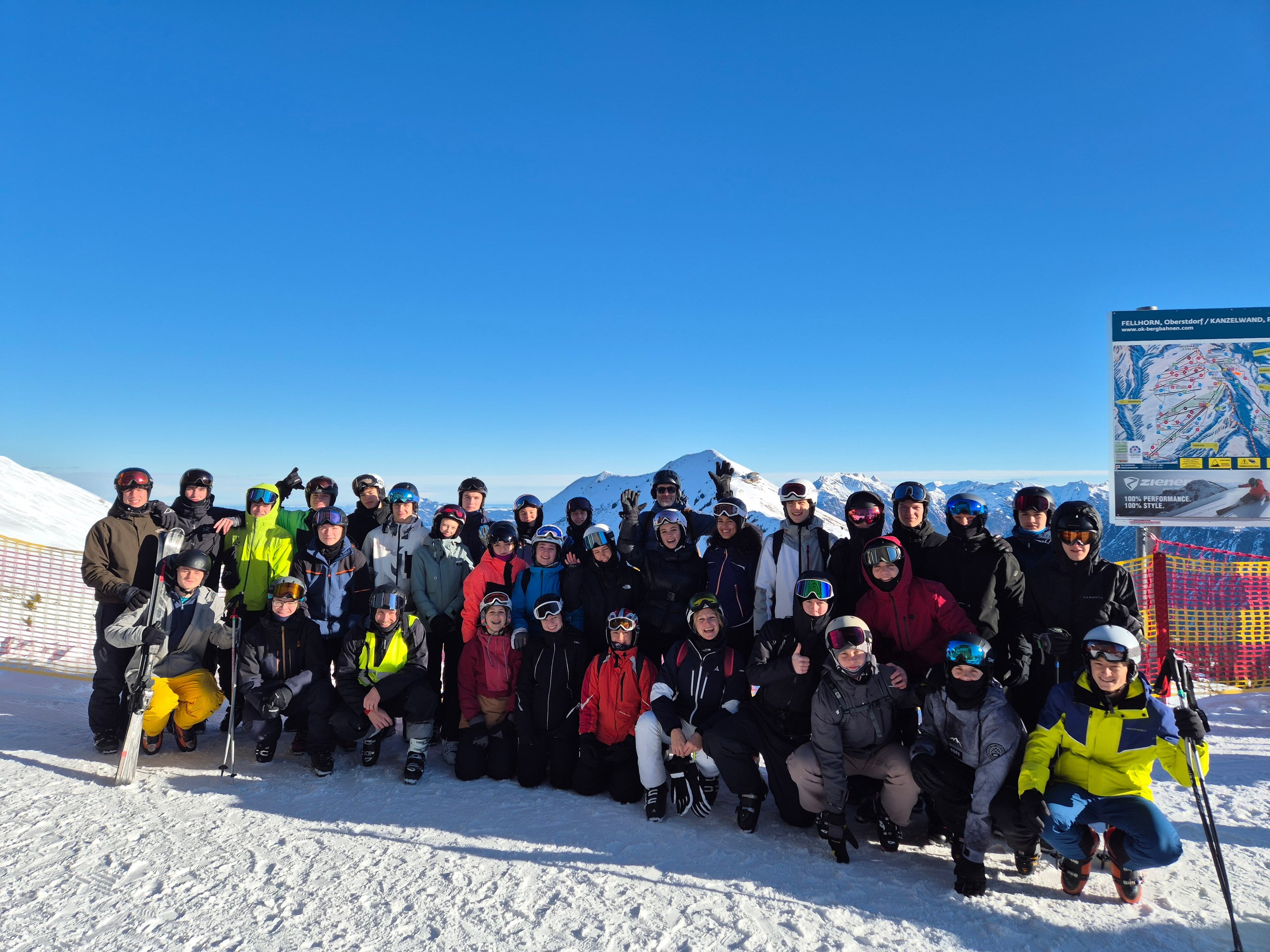 A group of people in ski gear poses on a snowy mountain against a clear blue sky with distant peaks.