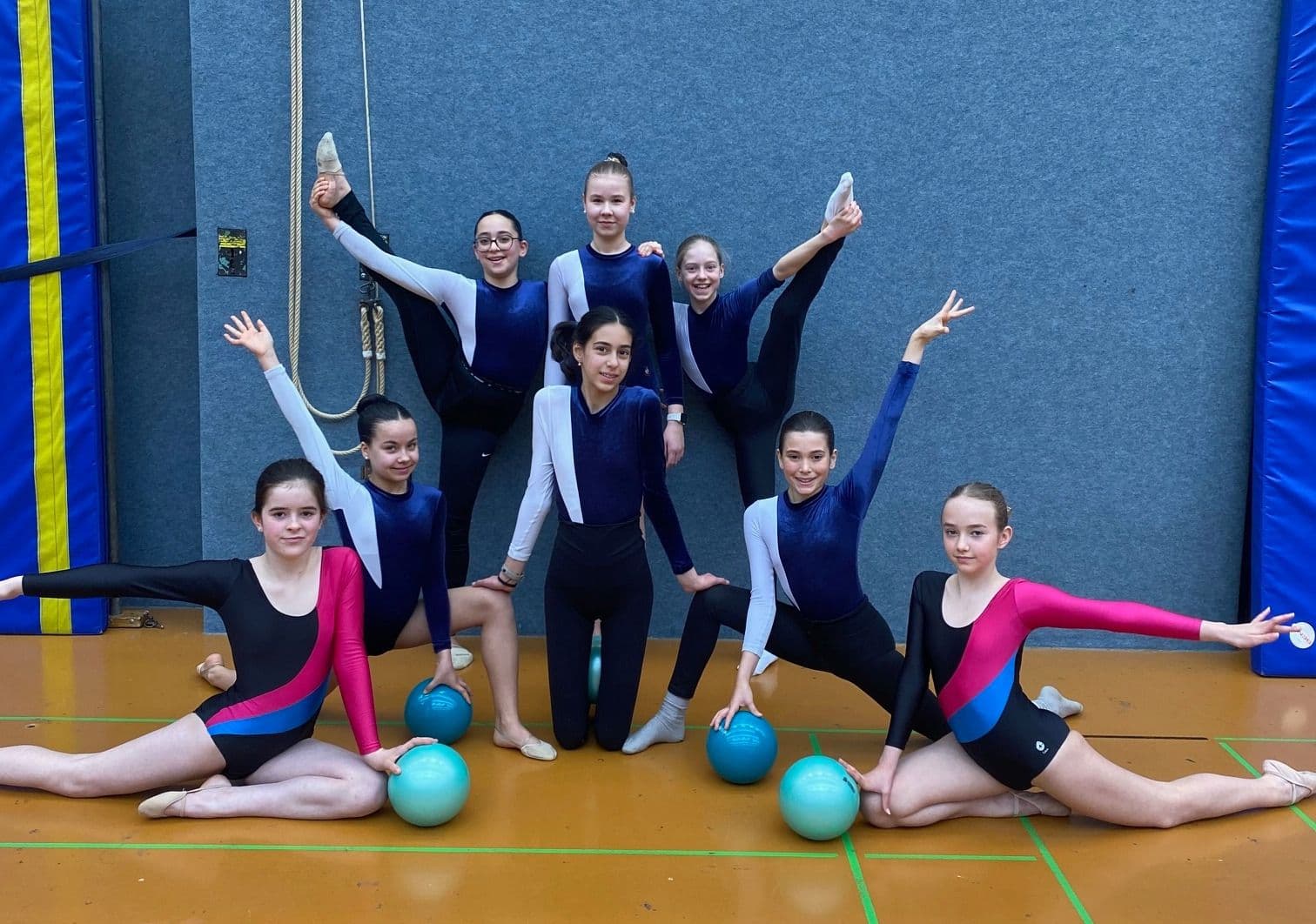 Eight young female gymnasts pose in a gym, several holding light blue balls.