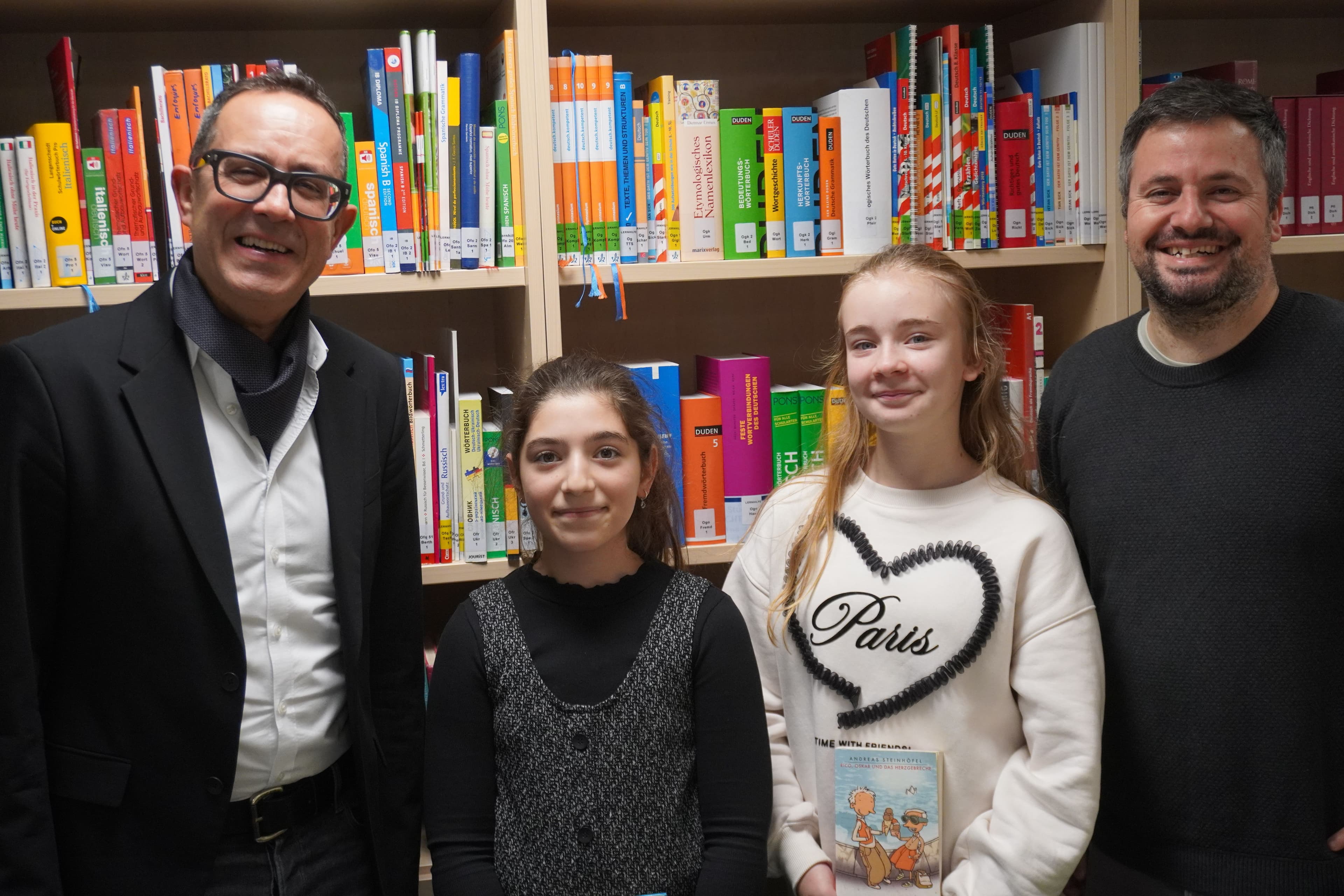 Two men and two girls smiling in front of bookshelves, one girl holding a book.