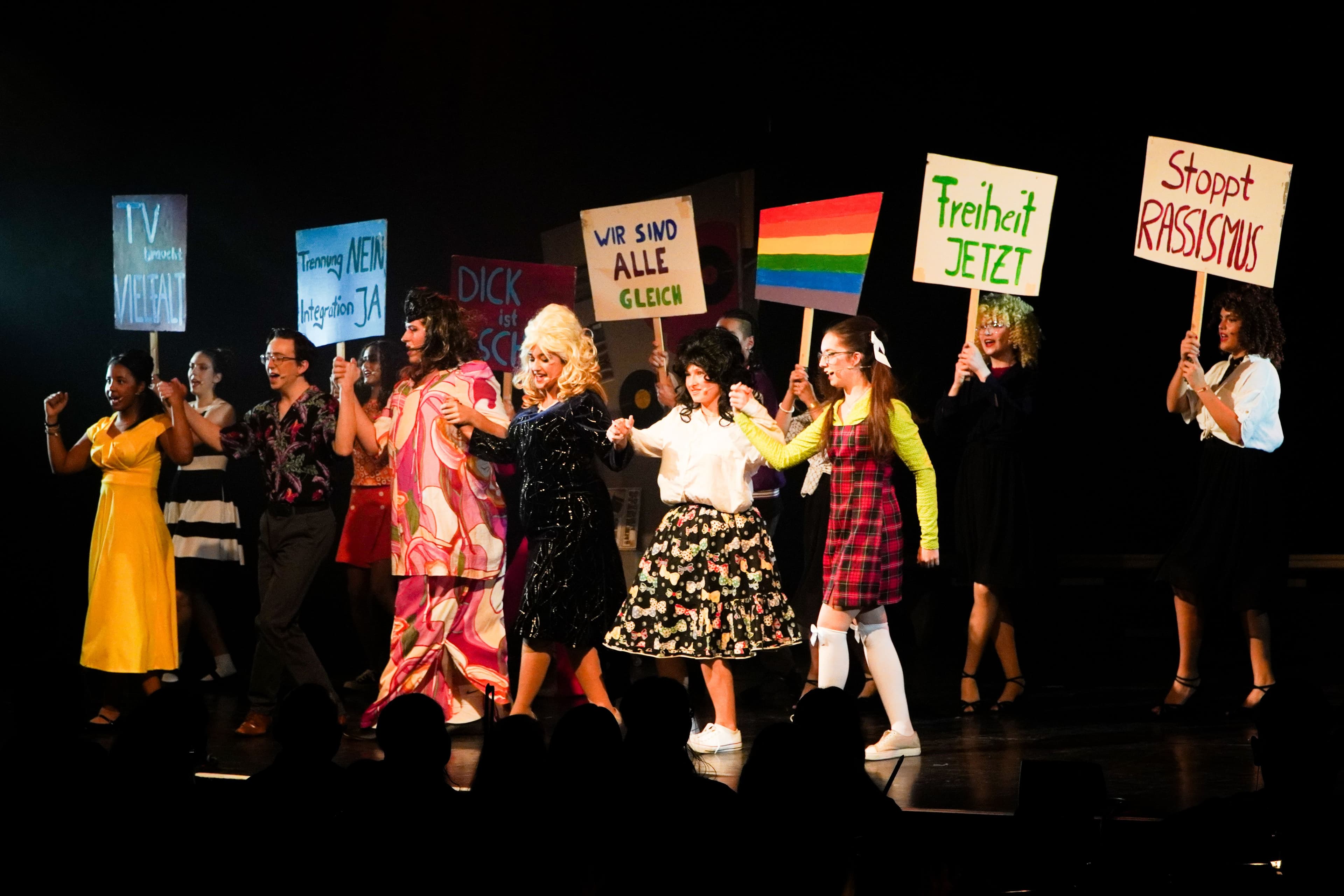 Schauspieler tanzen und demonstrieren auf einer Bühne, halten Schilder hoch.