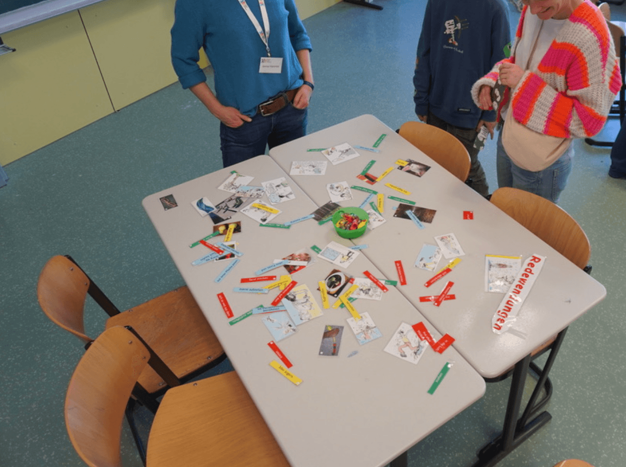 Three people stand around a classroom table covered with cards and colorful labels, suggesting an educational game.