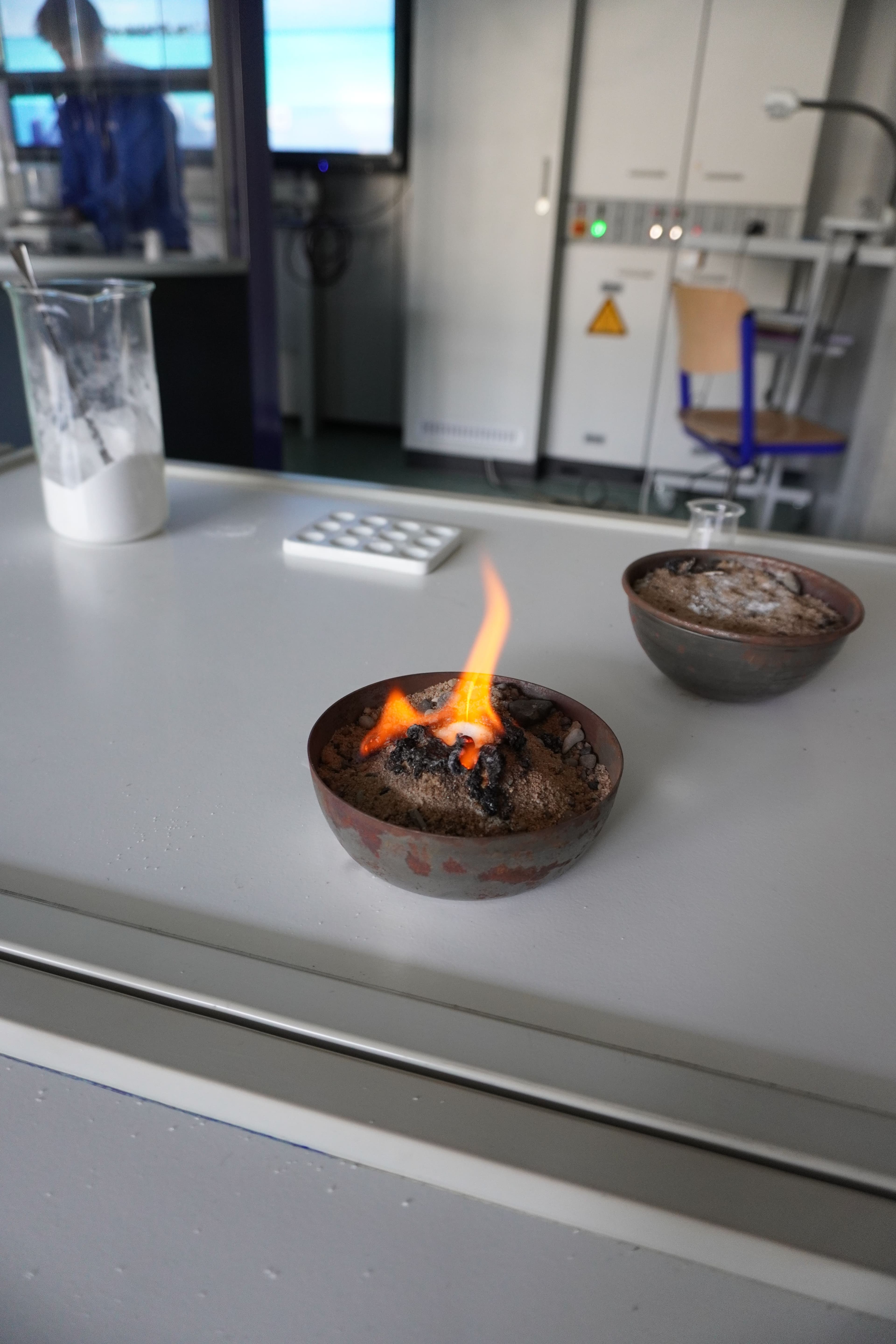 A metal bowl with a small orange flame burning in sand, next to another bowl of sand and a beaker of white powder, in a lab.