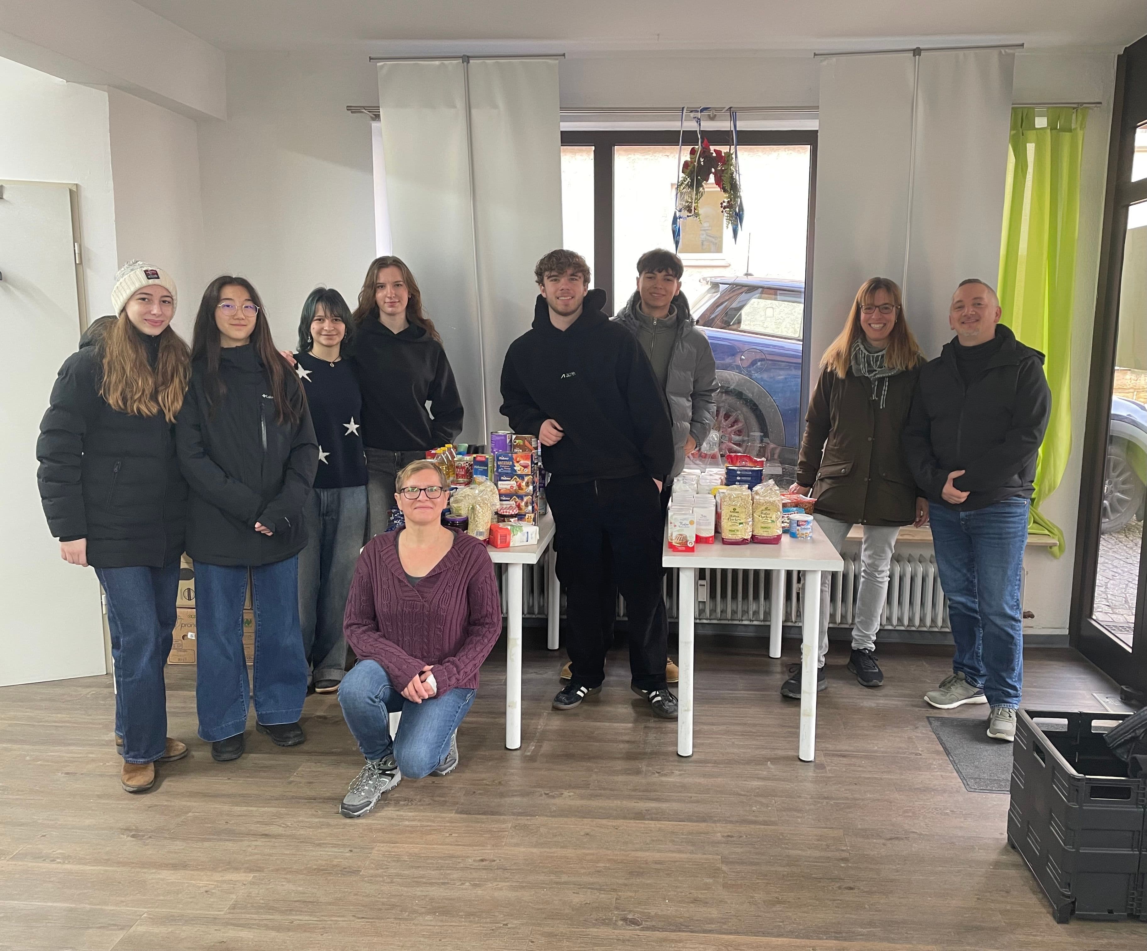Ten people, including young adults, stand with tables of groceries at a food distribution event.