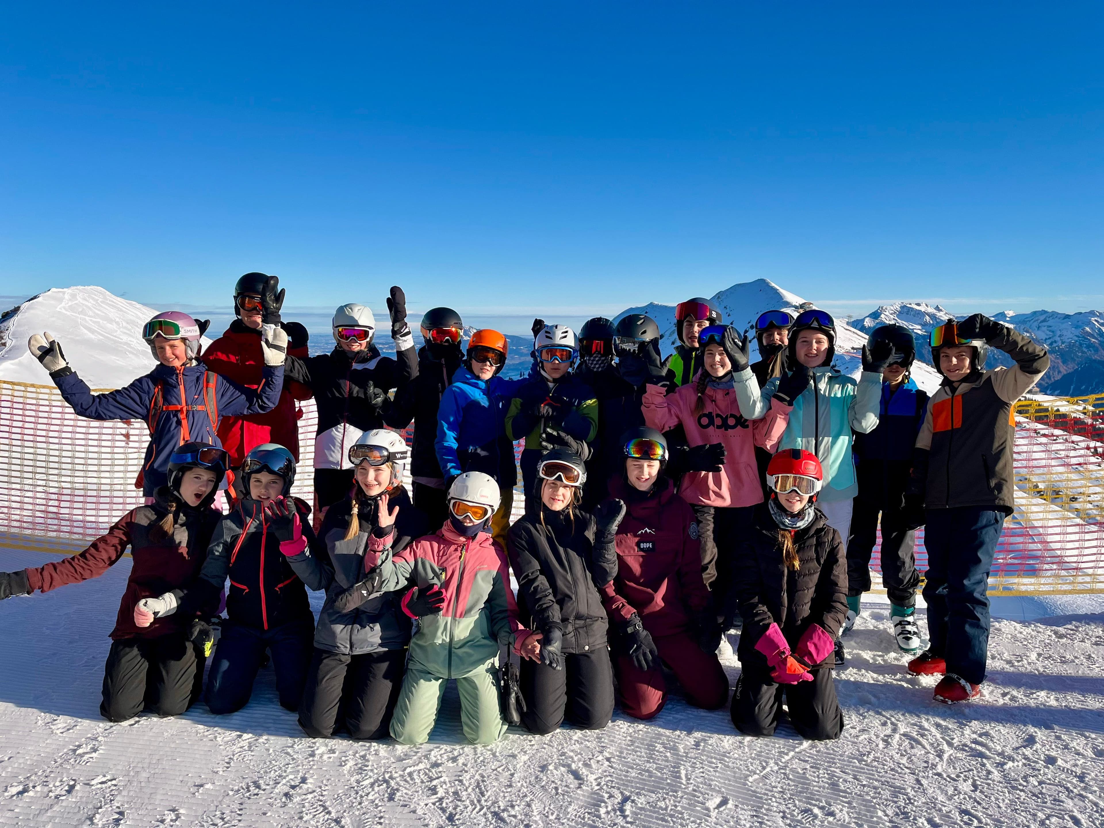 A group of young people in ski gear posing on a snowy mountain with clear blue sky and snow-capped peaks in the background.