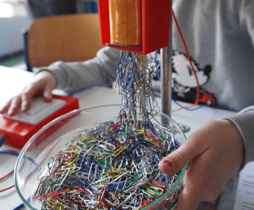 A child's hands conduct an electromagnet experiment, with many colorful paper clips clinging to an orange electromagnet above a bowl.
