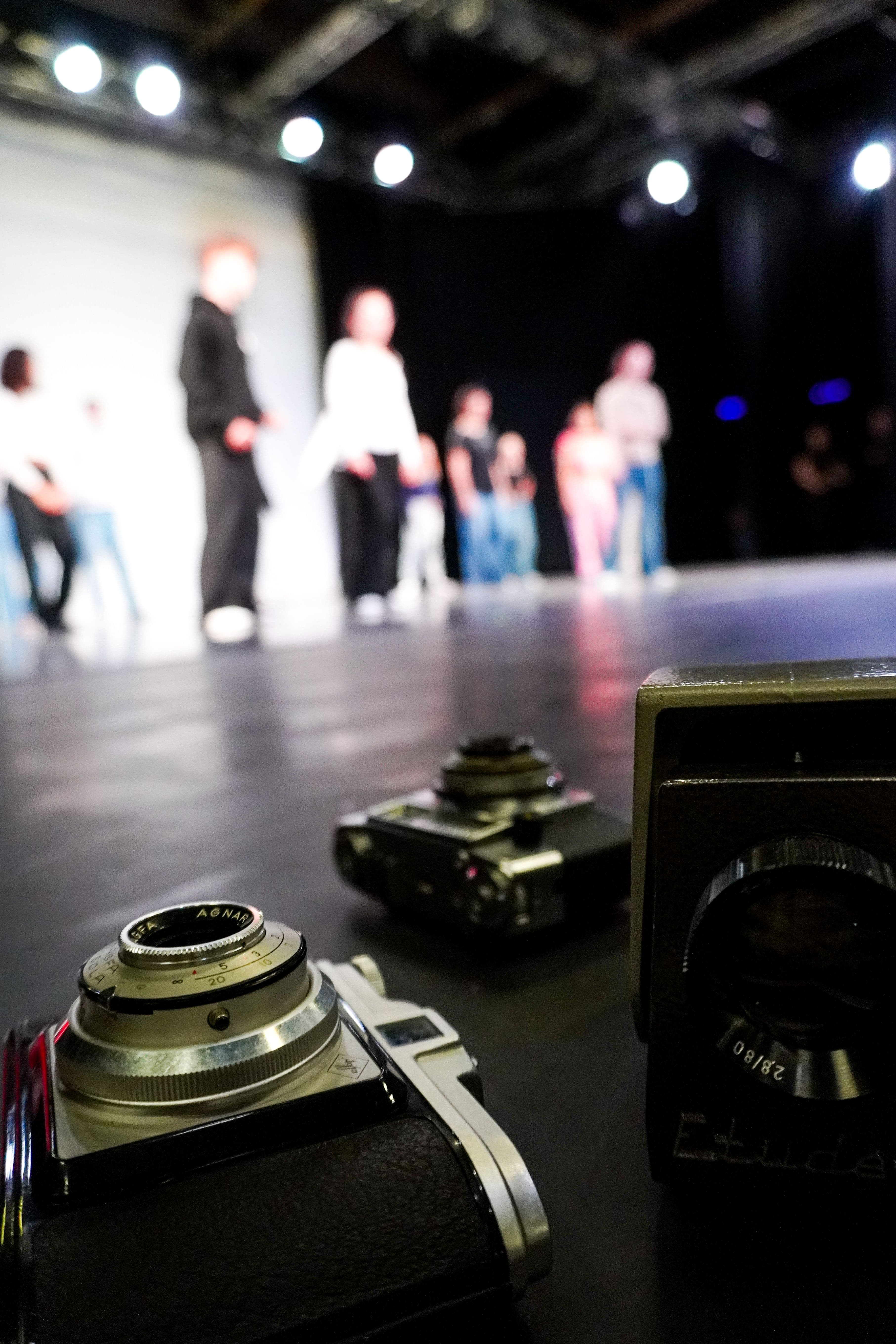Im Vordergrund eine alte Fotokamera, im Hintergrund eine Bühne mit Schauspielern, die proben.