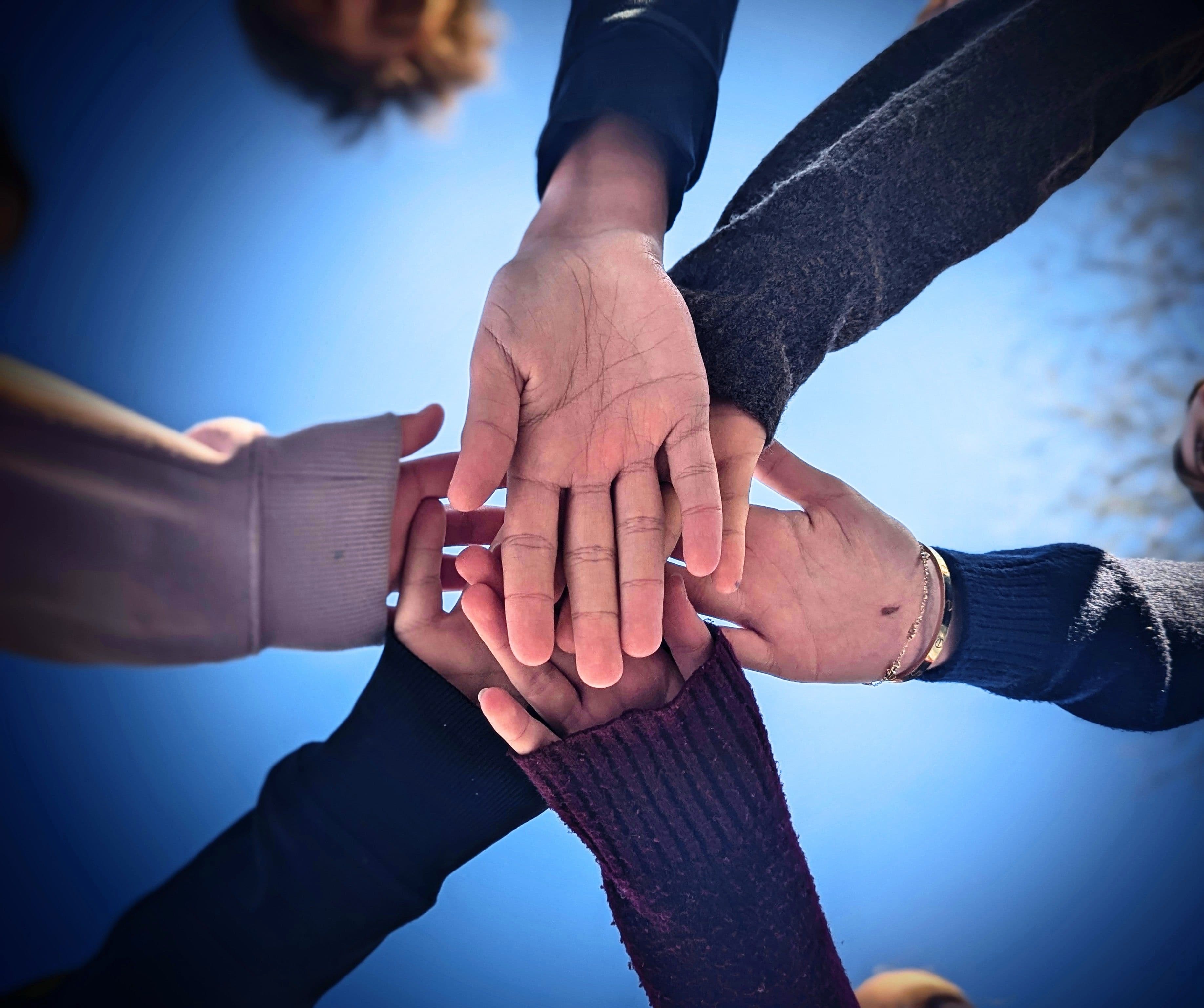 View from below of multiple hands stacked together against a bright blue sky.