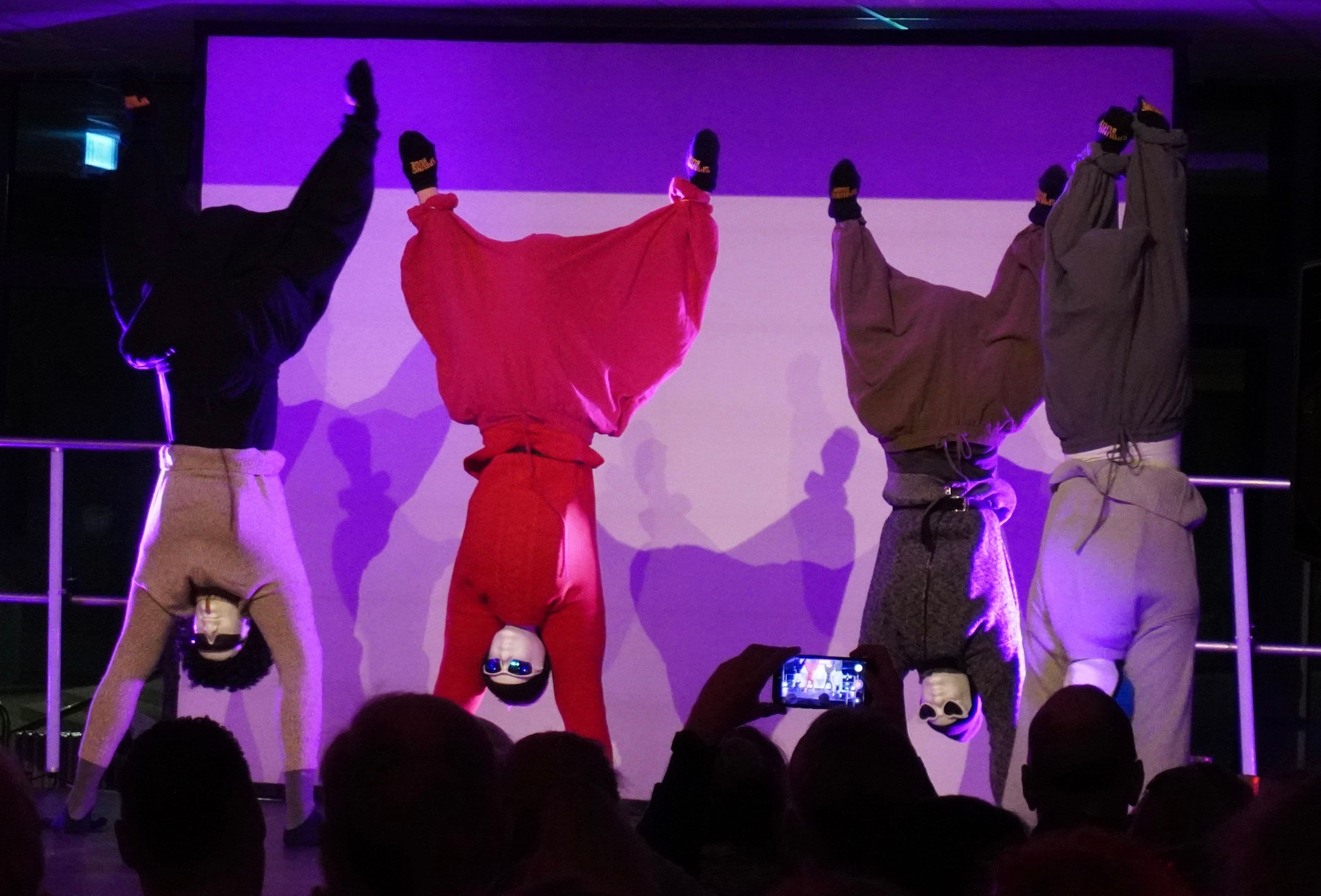 Four figures in colorful clothes and sunglasses perform handstands on a stage, viewed by an audience.