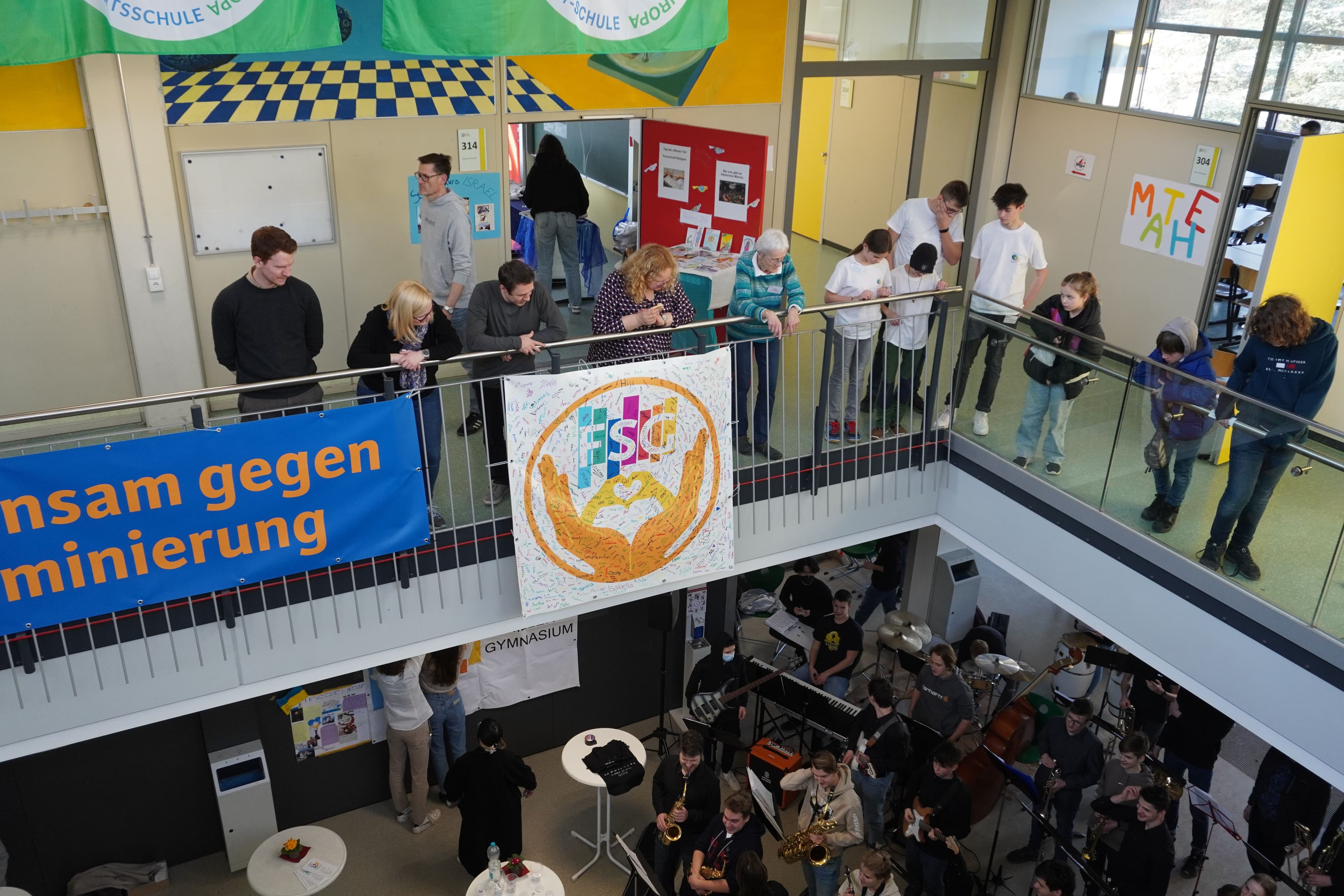 A multi-level school atrium where a student orchestra performs on a lower level, watched by students and adults from the upper balconies, with anti-discrimination banners displayed.