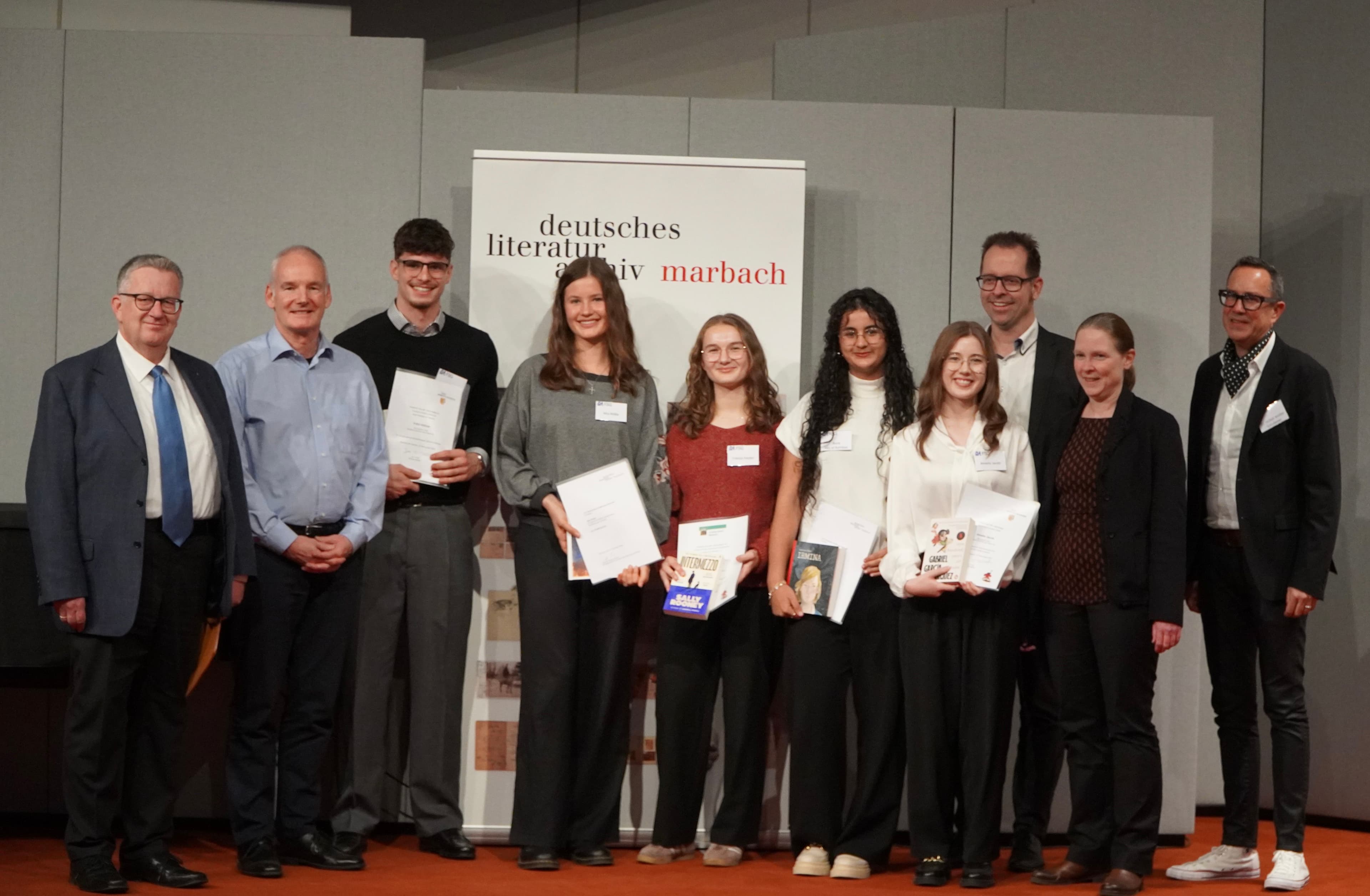 Ten people, a mix of adults and younger individuals, hold awards in front of a Deutsches Literaturarchiv Marbach banner.
