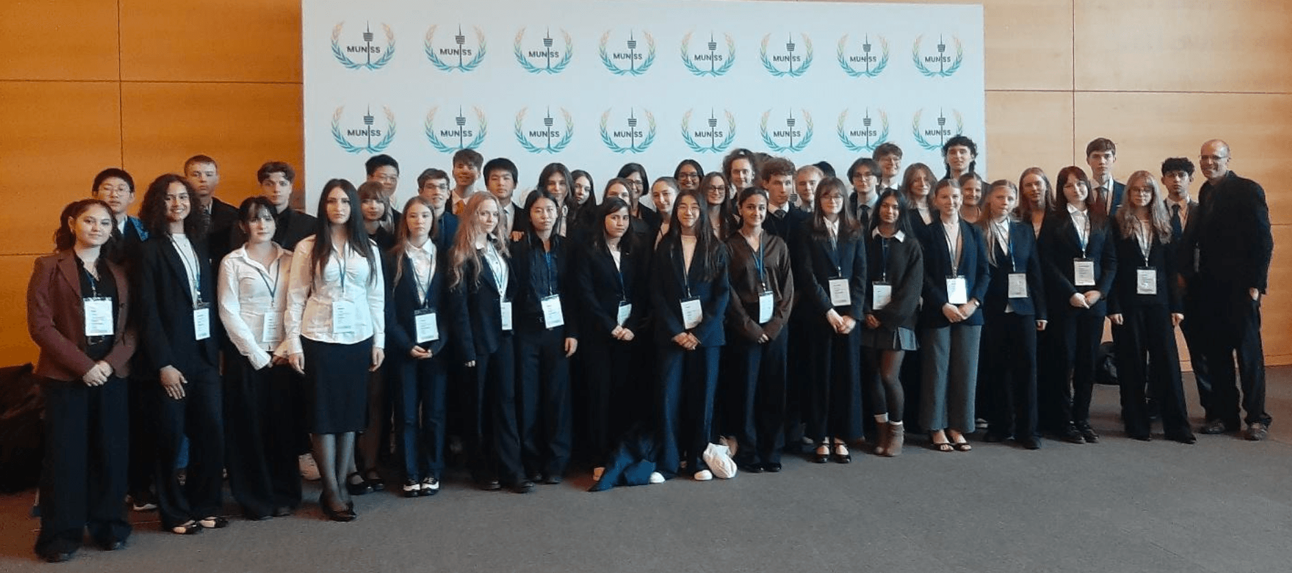 A large group of students and an adult in professional attire with ID badges pose in front of a wall with repeating logos.