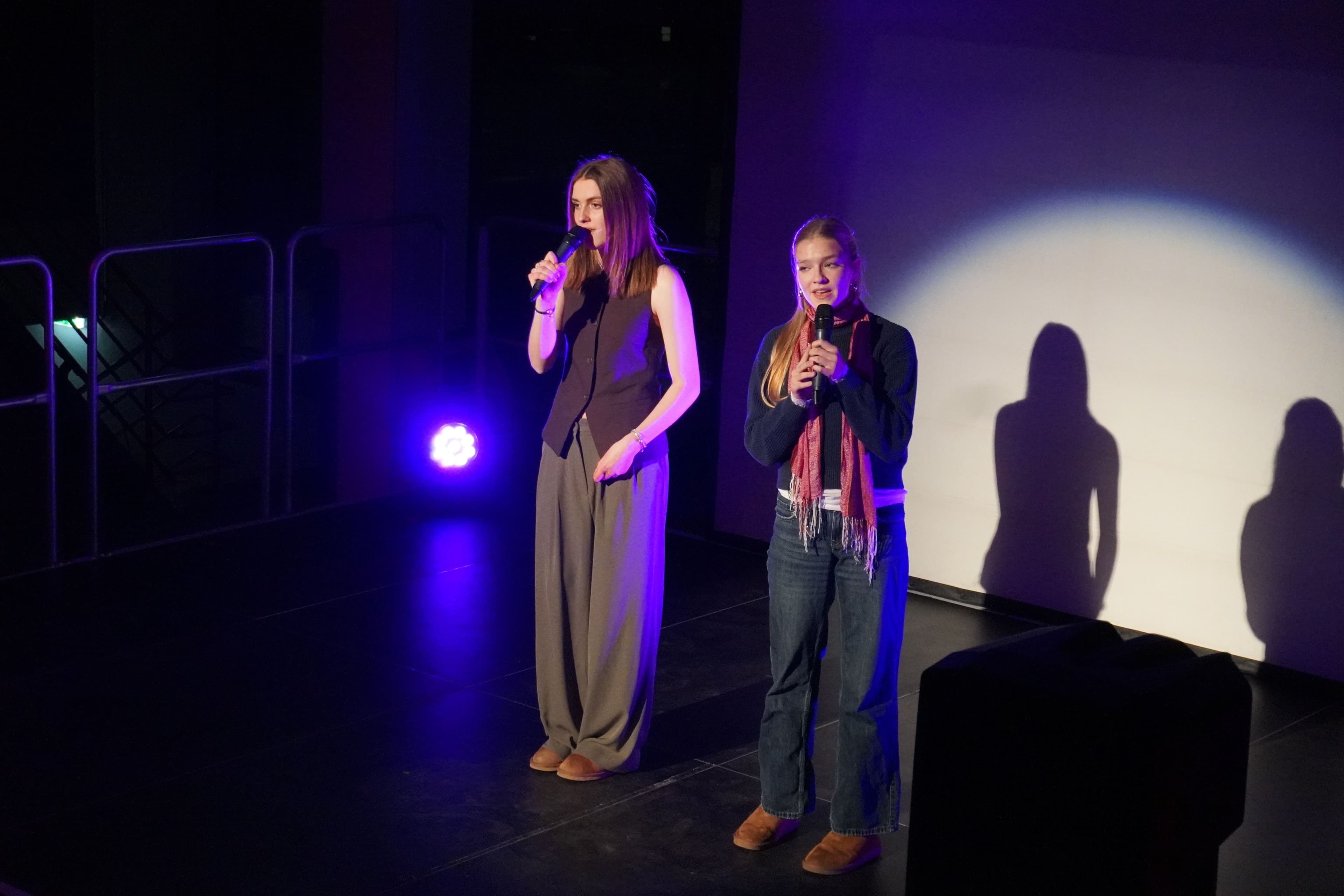 Two young women, each holding a microphone, stand on a dimly lit stage with purple lights and their shadows cast on a white wall.