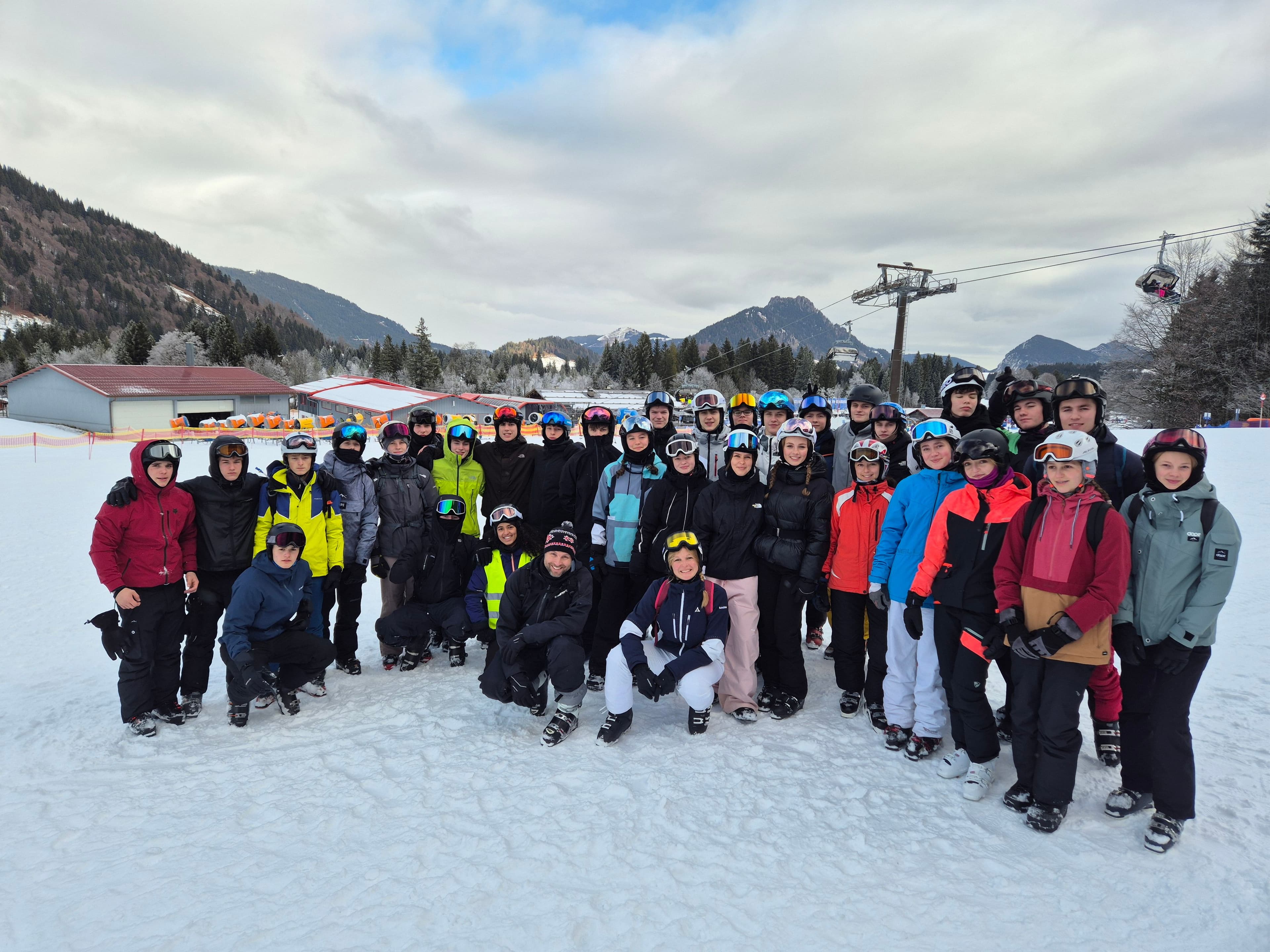 A large group of people in ski gear and helmets stands on a snowy mountain with a ski lift in the background.