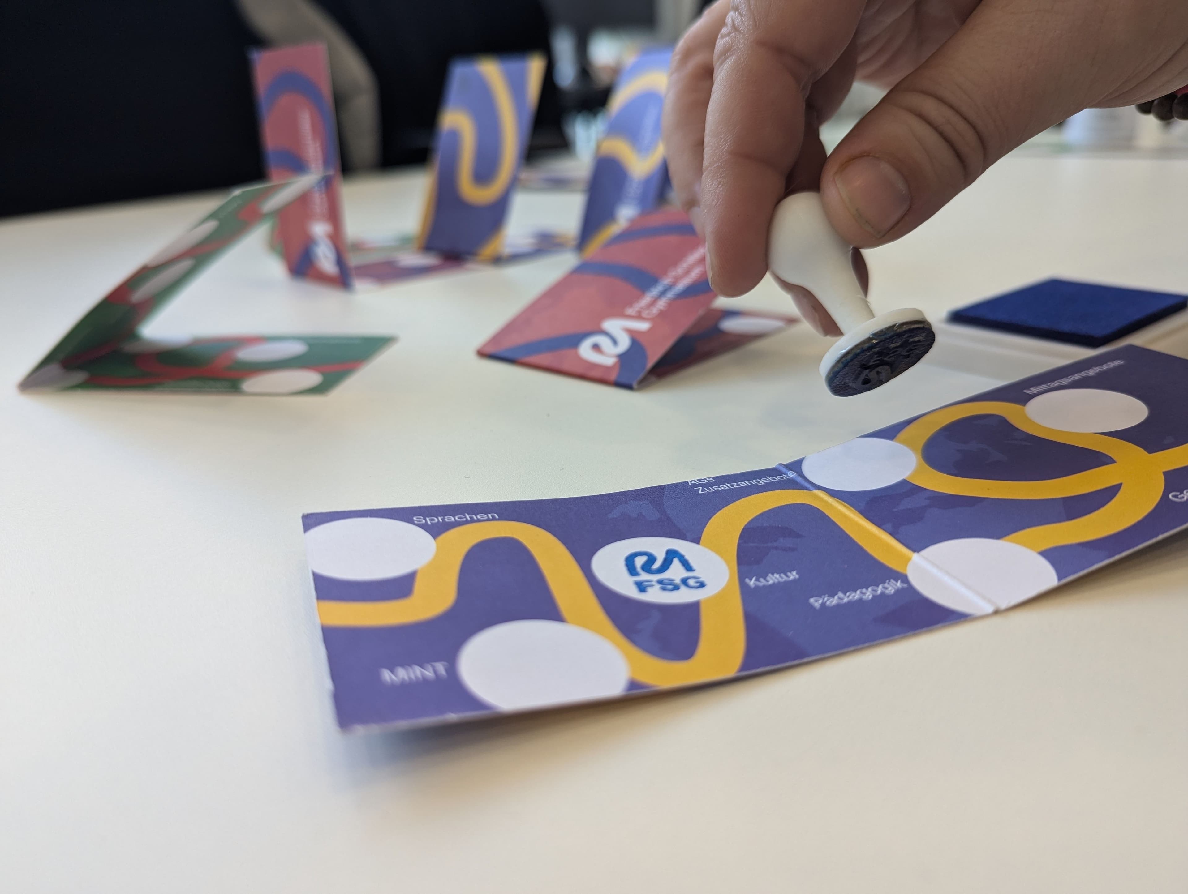 A hand holds a stamp over a purple folded paper strip with a yellow path and white circles, laid out on a white table with other colorful strips.