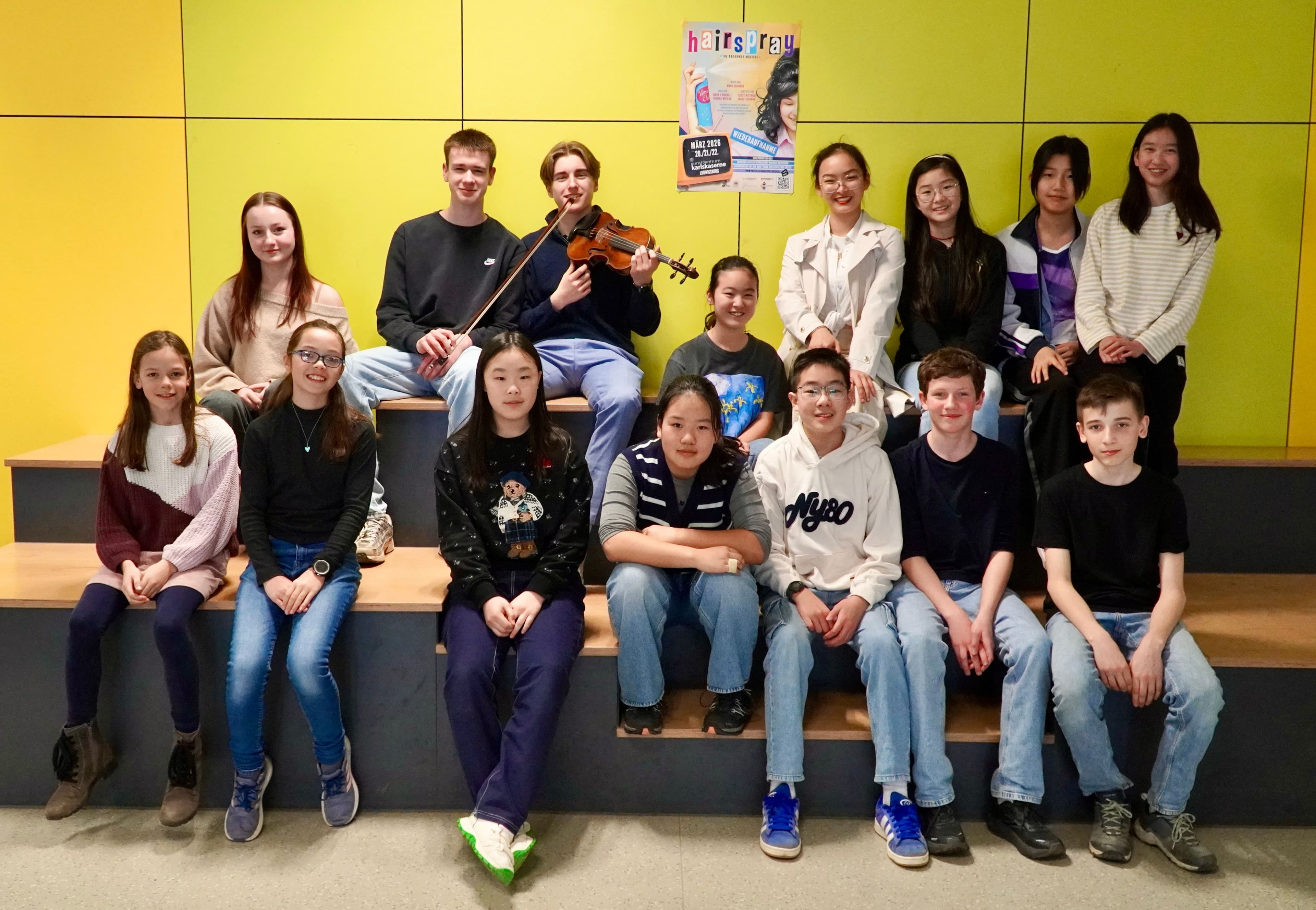 A diverse group of 17 teenagers sits on steps against a yellow wall, with one boy holding a violin.
