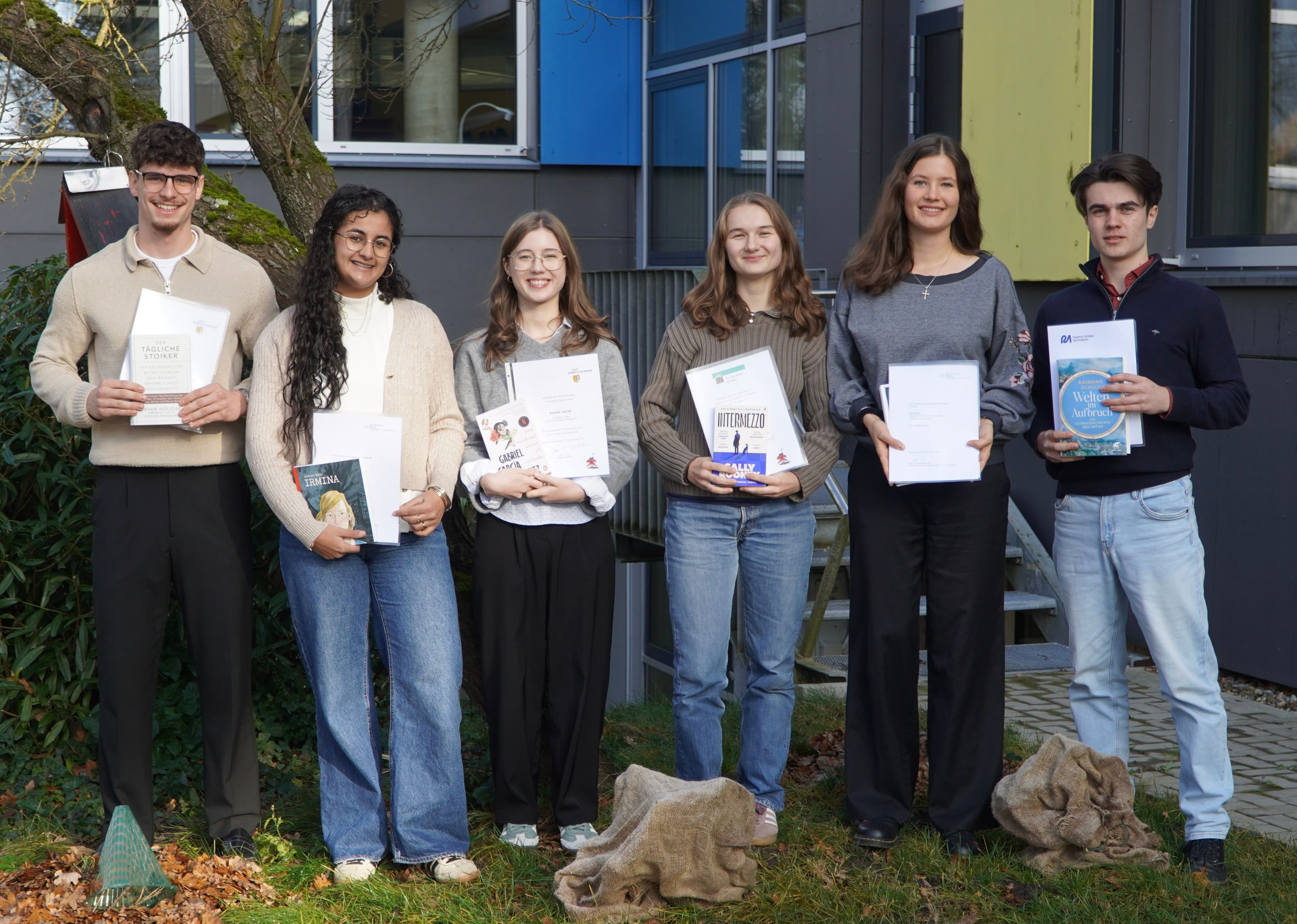Six smiling young adults, holding certificates and books, stand outdoors.