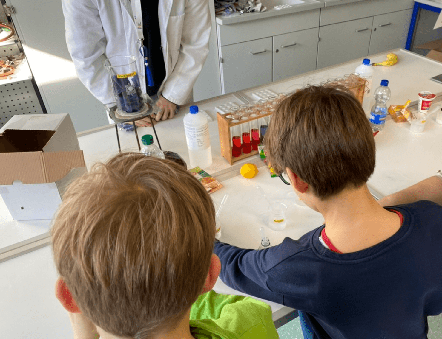 Two children at a lab table performing a science experiment, overseen by an adult in a lab coat.