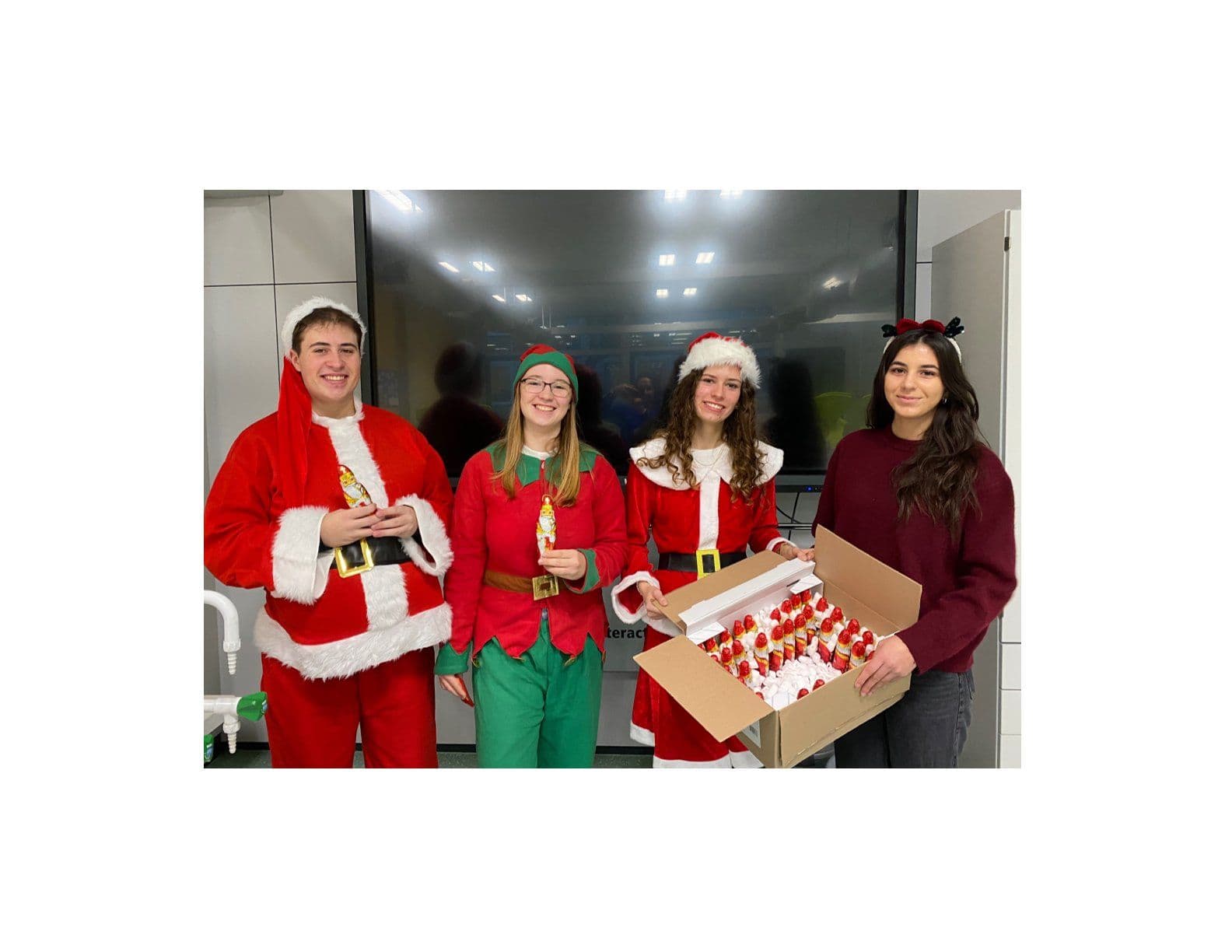 Four young people in Christmas outfits, holding festive figures and a box of Santa treats.