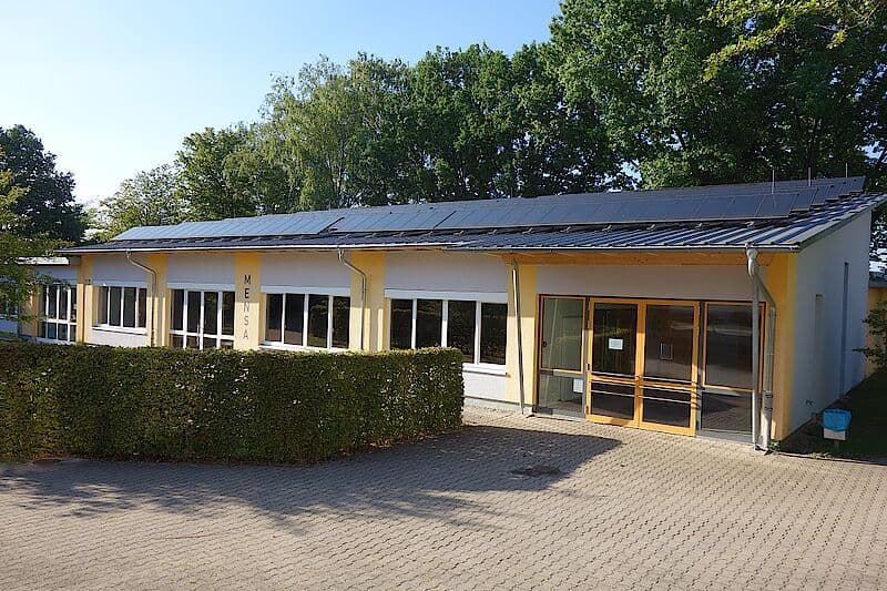 A modern building with solar panels on its roof, a hedge in front, and the word "MENSA" written vertically on the wall.