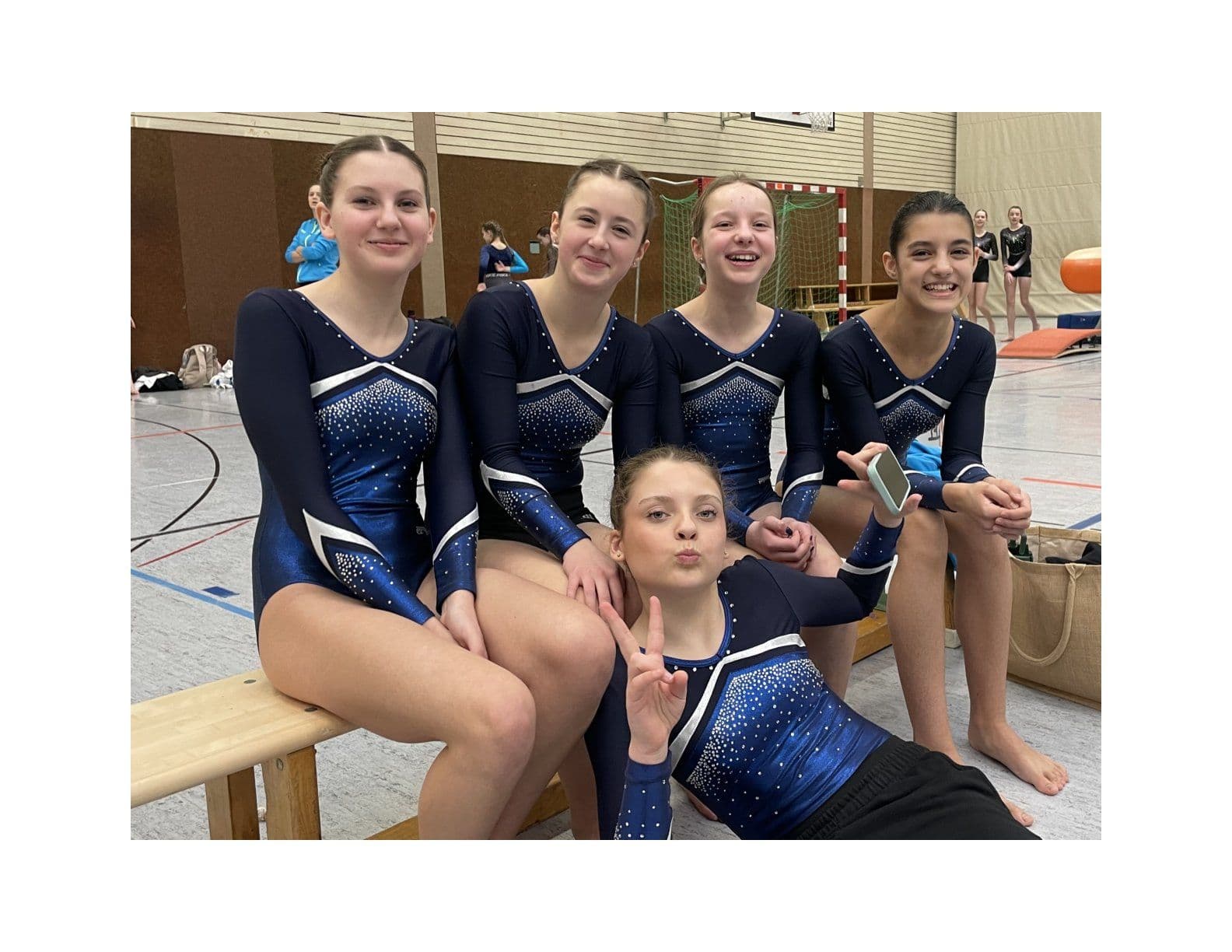 Five female gymnasts in blue leotards smiling and posing happily in a gym.