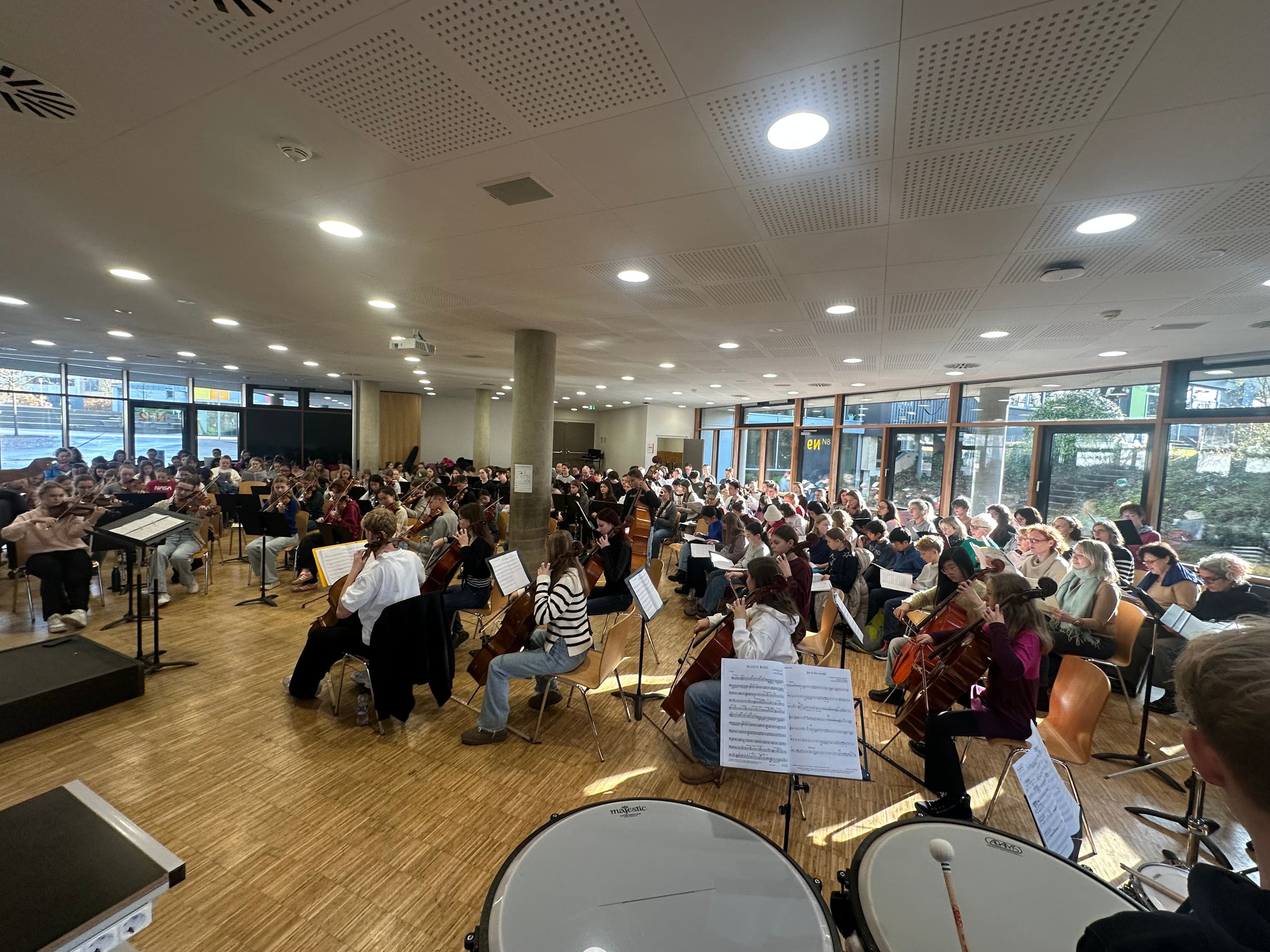 A student orchestra rehearsing string instruments and percussion in a bright room.